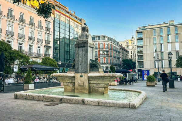 Fountain at Plaza de la Luna, surrounded by modern and historic buildings in Madrid, Spain, under a clear blue sky.