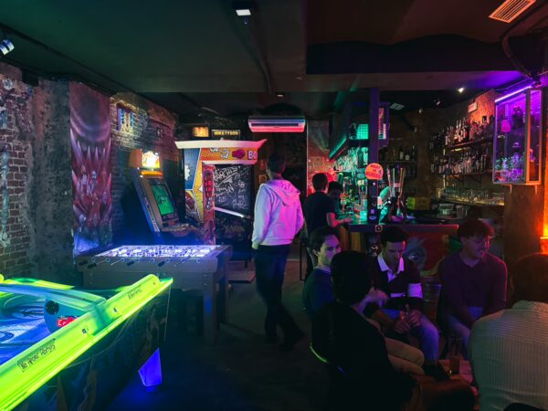 Vibrant arcade scene at Next Level Bar in Madrid, featuring neon lights, pinball machines, and lively patrons enjoying the atmosphere.