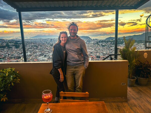 Couple posing against a vibrant sunset over Quito, with cityscape and a drink on the table in the foreground.