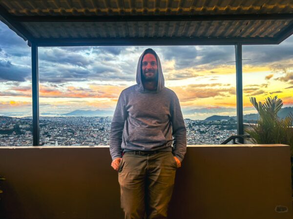Traveler in a hoodie stands against a backdrop of a colorful sunset over Quito, Ecuador, with cityscape and mountains visible.