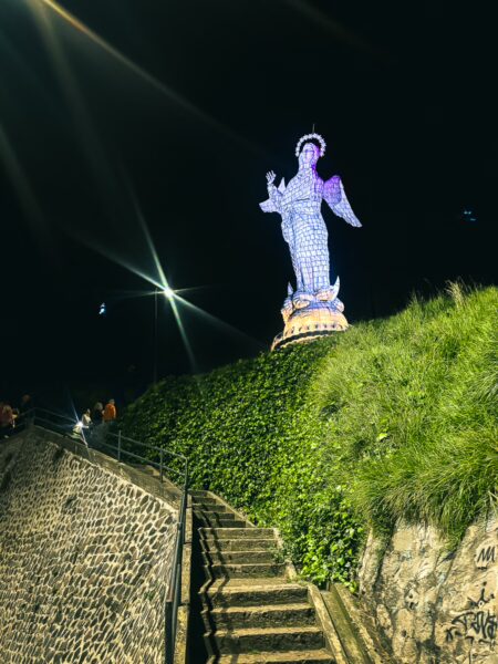 Glowing statue of the Virgin Mary atop a hill surrounded by lush greenery, with stairs leading up in the night in Quito, Ecuador.