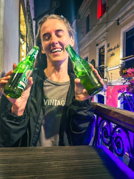 A woman enjoys two green bottles of beer, smiling at a lively outdoor bar in La Ronda, Quito, Ecuador.