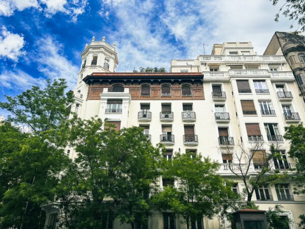 Elegant white buildings with balconies and a tower stand amidst lush green trees under a blue sky in Madrid, Spain.