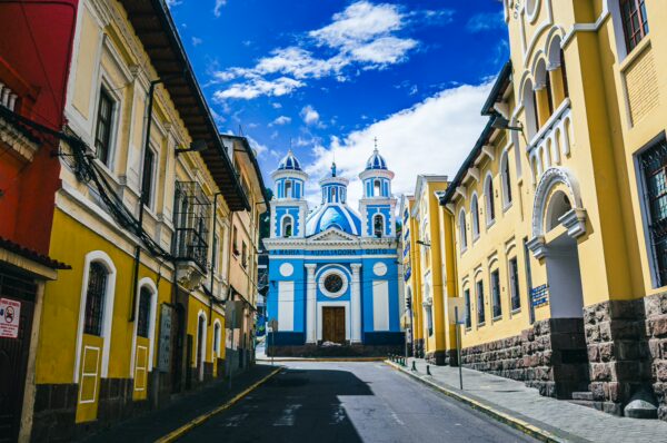 Vibrant blue Templo Maria Auxiliadora de La Tola stands at the end of a cobblestone street in Quito, Ecuador, framed by colorful buildings.