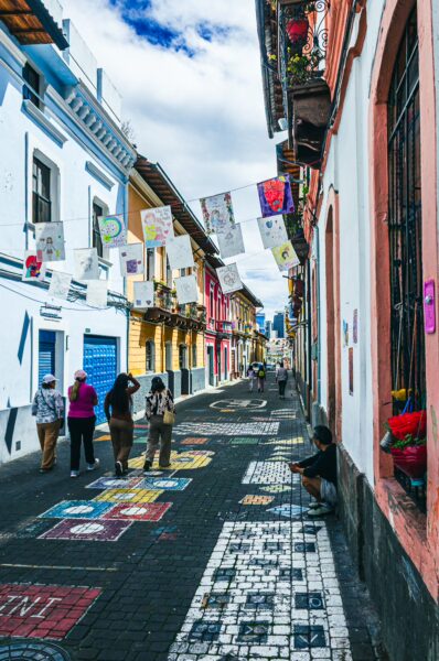 Colorful papel picado flutters above Vicente León Street in Quito, Ecuador, as visitors explore vibrant murals and charming buildings.
