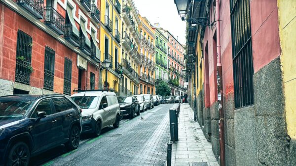 Colorful buildings line a narrow street in Madrid, Spain, with parked cars and a hint of greenery in the background.