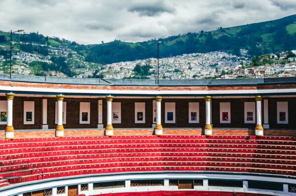 Vibrant red seating surrounds the arena at Plaza de Toros Belmonte, with green hills and Quito's skyline visible in the background.
