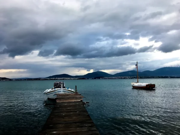 Calm waters reflect a cloudy sky as two boats rest by a wooden pier in Chalkis, creating a serene coastal scene.