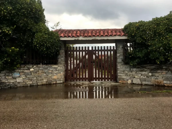 Charming stone entrance with a wooden gate, framed by lush greenery, in Chalkis after a recent rainfall.