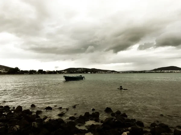 Calm waters in Chalkis feature a lone boat, surrounded by dark rocky shores under a moody, cloudy sky.