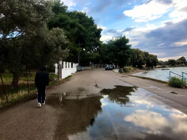 Walking along a tranquil waterfront path in Chalkis, reflections in puddles and lush greenery create a serene atmosphere.