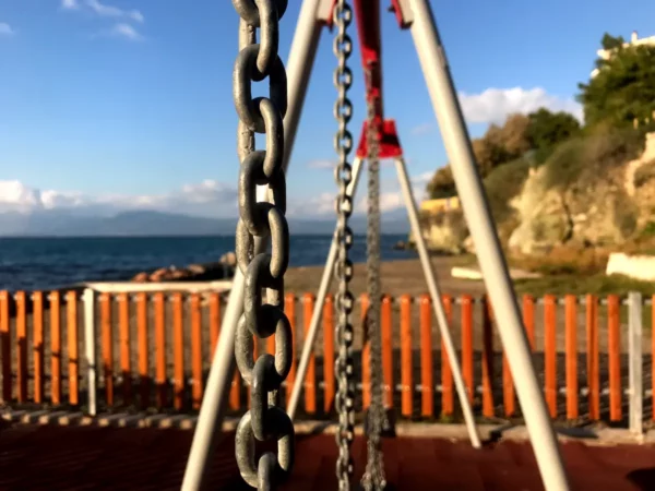 Rusty chains hang from a swing set overlooking the calm sea in Chalkis, framed by a vibrant orange fence and scenic cliffs.
