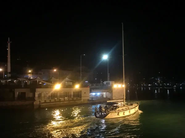 Sailboat gliding through illuminated waters at night, with waterfront buildings and reflections in Chalkis.