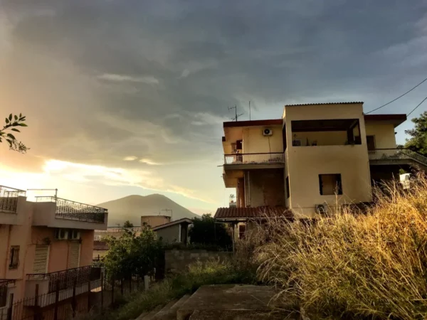 Chalkis scenery features a two-story house against a dramatic sky at sunset, with wild grass in the foreground and distant hills.