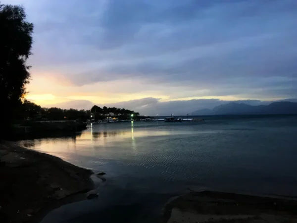 Serene twilight over Chalkis, with gentle waves reflecting the soft colors of sunset and a distant marina illuminated in the evening light.