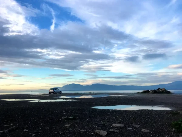 Calm waters and scattered clouds reflect the serene landscape near Chalkis, with a lone car parked by the shoreline.