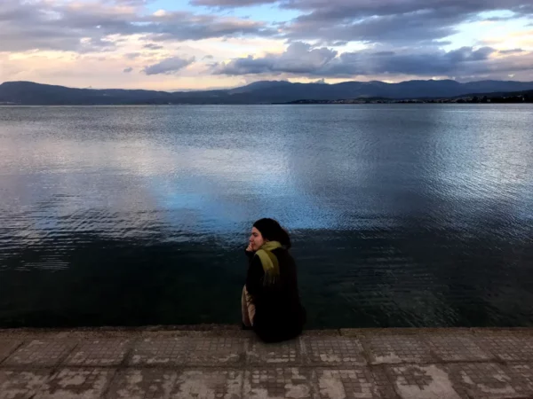 Seated by the water's edge, a woman enjoys a peaceful moment overlooking the calm lake in Chalkis, with mountains in the distance.