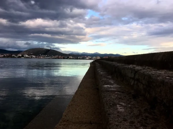 Serene waterfront view in Chalkis, showcasing a stone pier, calm water, and dramatic cloud formations above distant mountains.