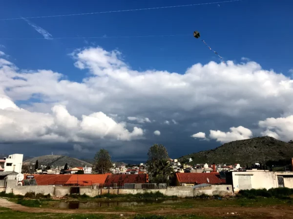 Colorful kite flying high against a cloudy sky above Chalkis, showcasing a vibrant landscape with nearby rooftops and hills.