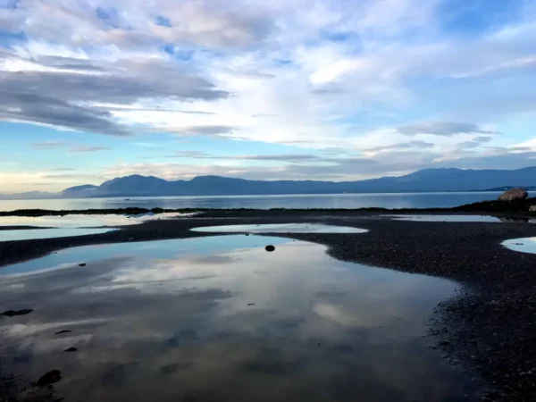 Calm waters reflecting the sky and mountains near Chalkis, with patches of gravel creating a serene coastal landscape.