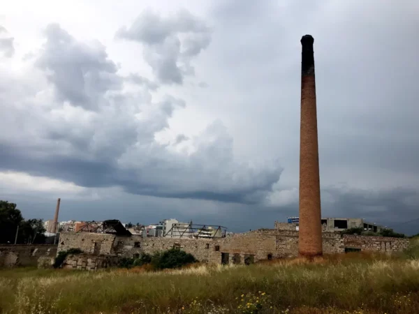 Weathered brick chimney stands tall among green grass and ruins under a cloudy sky in Chalkis.