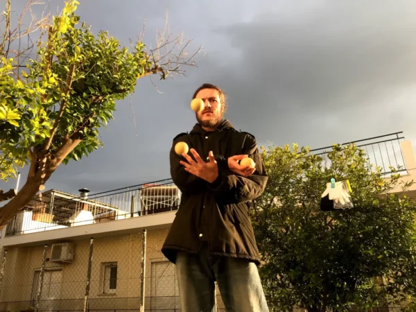 Juggler skillfully tossing three oranges under overcast skies in Chalkis, with trees and a building in the background.