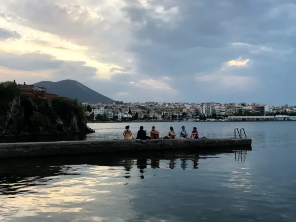 Sunset over the calm waters of Chalkis, with people seated on a pier and colorful buildings reflecting in the water.