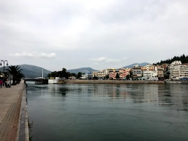 Chalkis waterfront features calm waters reflecting colorful buildings along the shore, with mountains in the background under a cloudy sky.