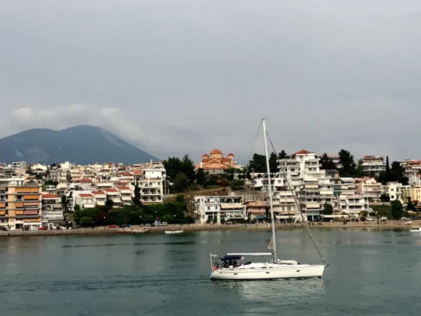 Sailboat gliding across the calm waters with Chalkis' colorful buildings and mountains in the background under a cloudy sky.