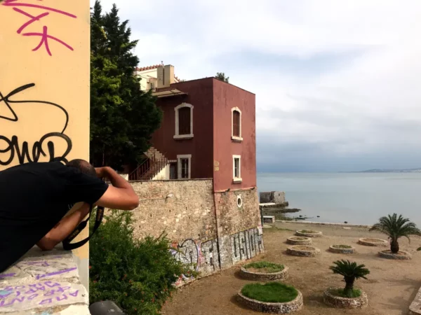 Chalkis features a photographer capturing the scenic shoreline, with a pink building, palm trees, and calm waters in the background.
