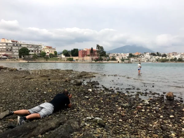 A photographer captures a person standing in the shallow waters of Chalkis, with rocky shorelines and coastal buildings in the background.