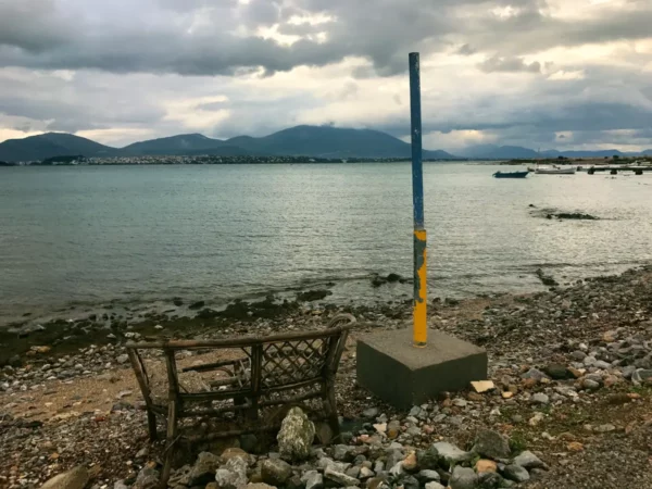 Rustic bench near the shoreline, with calm waters and distant mountains under a cloudy sky in Chalkis.