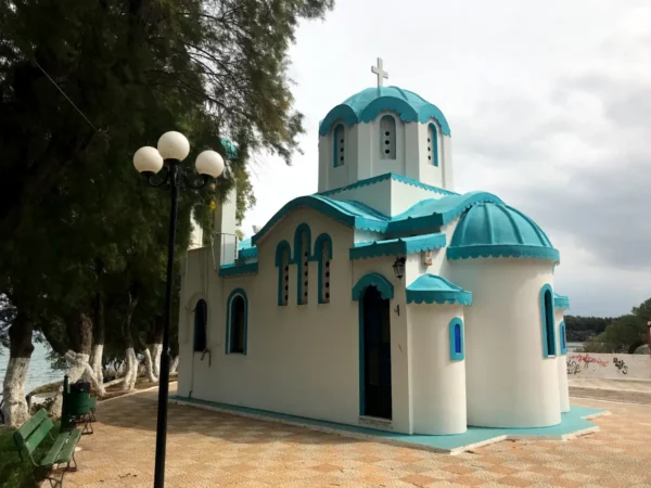 Charming white and turquoise chapel with domed roof, surrounded by trees and a checkered pavement in Chalkis.