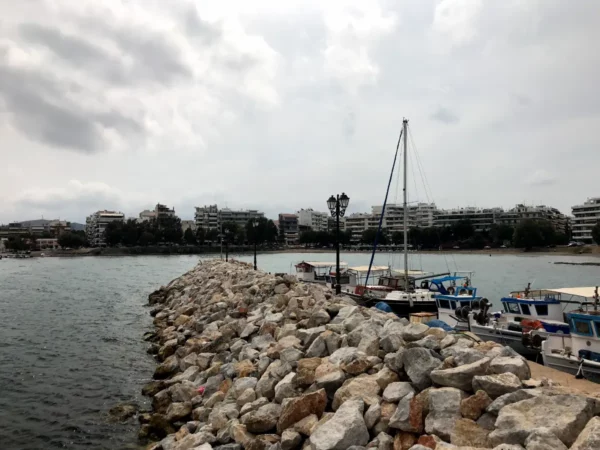 Chalkis waterfront features a rocky pier leading to moored boats with modern buildings lining the horizon under a cloudy sky.