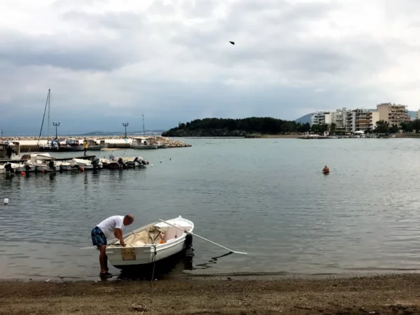 A man preparing a small boat on the sandy shore of Chalkis, surrounded by calm waters and distant buildings under a cloudy sky.