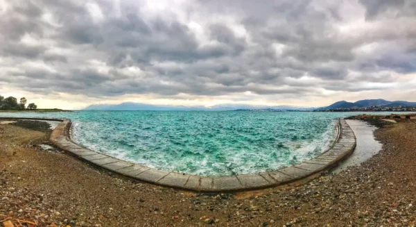 Turquoise waters lap against a pebbled shore, under dramatic gray skies in Chalkis, showcasing a serene coastal scene.