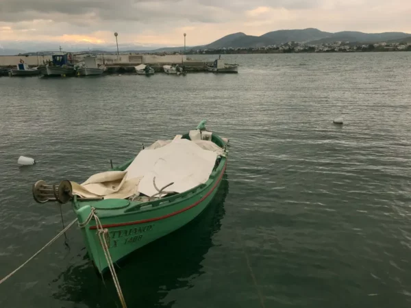 Green fishing boat moored in calm waters under overcast skies, with distant hills and a small harbor in Chalkis.