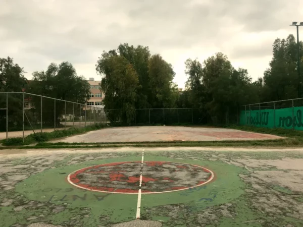 Faded basketball court with vibrant graffiti, surrounded by trees, in Chalkis, creating a nostalgic urban scene.