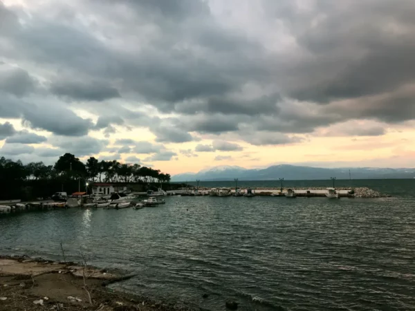 Chalkis waterfront at dusk, featuring boats docked near a rocky pier, with soft waves and cloudy skies in the background.
