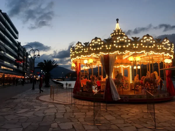Colorful carousel illuminated with lights at dusk, with palm trees and modern buildings lining the waterfront in Chalkis.