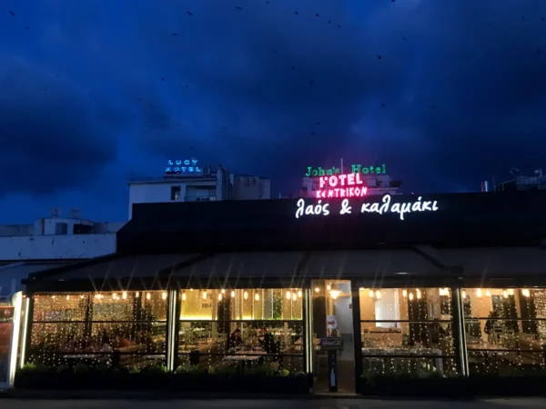 Brightly lit café with festive outdoor seating, under a moody sky in Chalkis, showcasing vibrant neon signage.