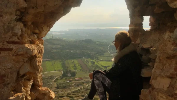 A woman sits by a stone window, gazing over lush green landscapes and shimmering water near Chalkis.