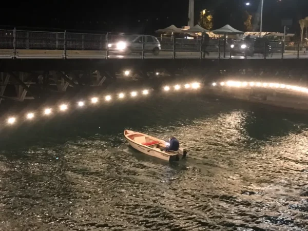A small boat navigates the illuminated waters under a bridge in Chalkis, with distant vehicles and vibrant reflections.