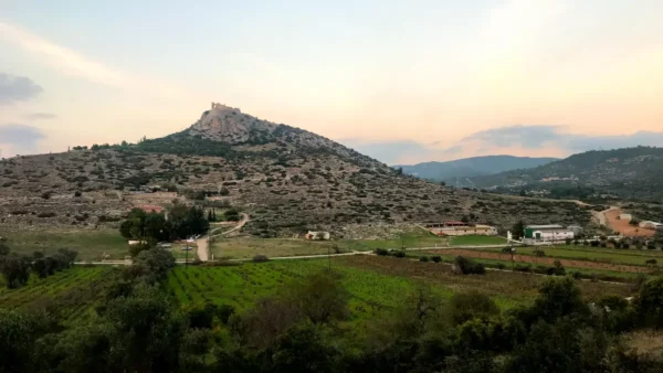 Rolling hills and scattered farms surround a distant hilltop castle, highlighting the natural beauty of Chalkis at sunset.
