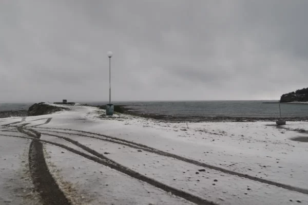 Snow-covered shore with tire tracks leading to the sea and a lone lamp post under a cloudy sky in Chalkis, Euboea, Greece.