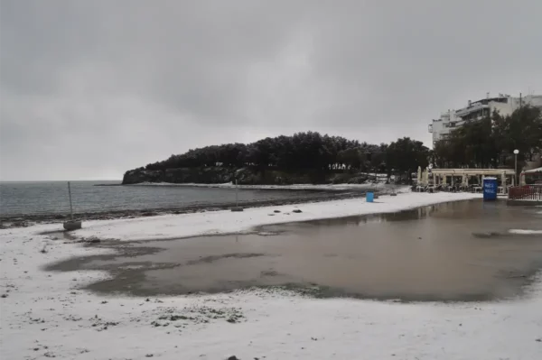 Chalkis beach in Euboea, Greece, features a snowy shoreline and a calm sea under a cloudy sky, with nearby trees and buildings.