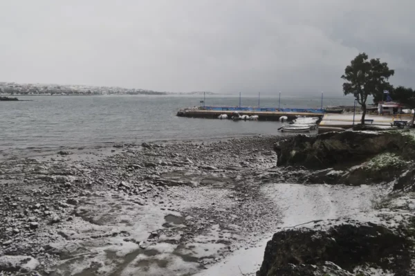 Snow-covered shoreline at Chalkis, Euboea, Greece, with a calm gray sea, rocky beach, and moored boats in the harbor.