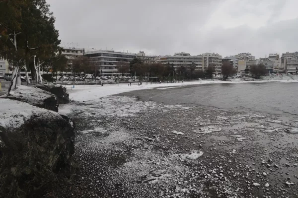 Snow-covered shoreline and rocky coast in Chalkis, Euboea, Greece, with grey skies and buildings lining the horizon.