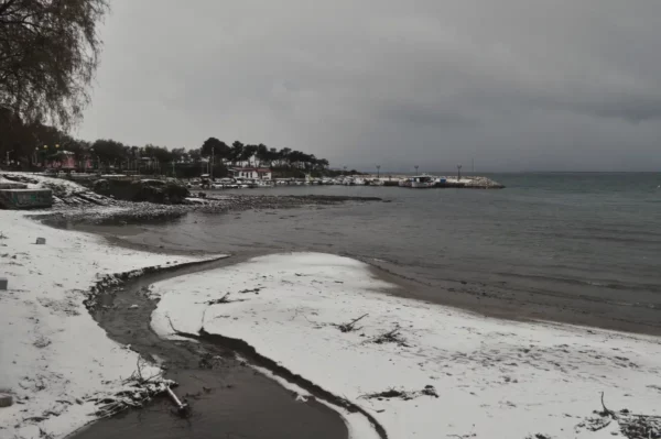 Snow-covered shoreline curves along the calm waters of Chalkis, Euboea, Greece, under a gray, overcast sky.