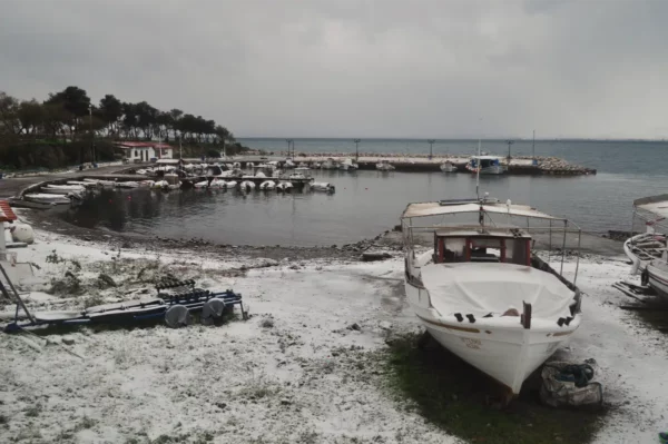 Snow-dusted shoreline with boats at rest near Chalkis, Euboea, Greece, framed by overcast skies and greenery.